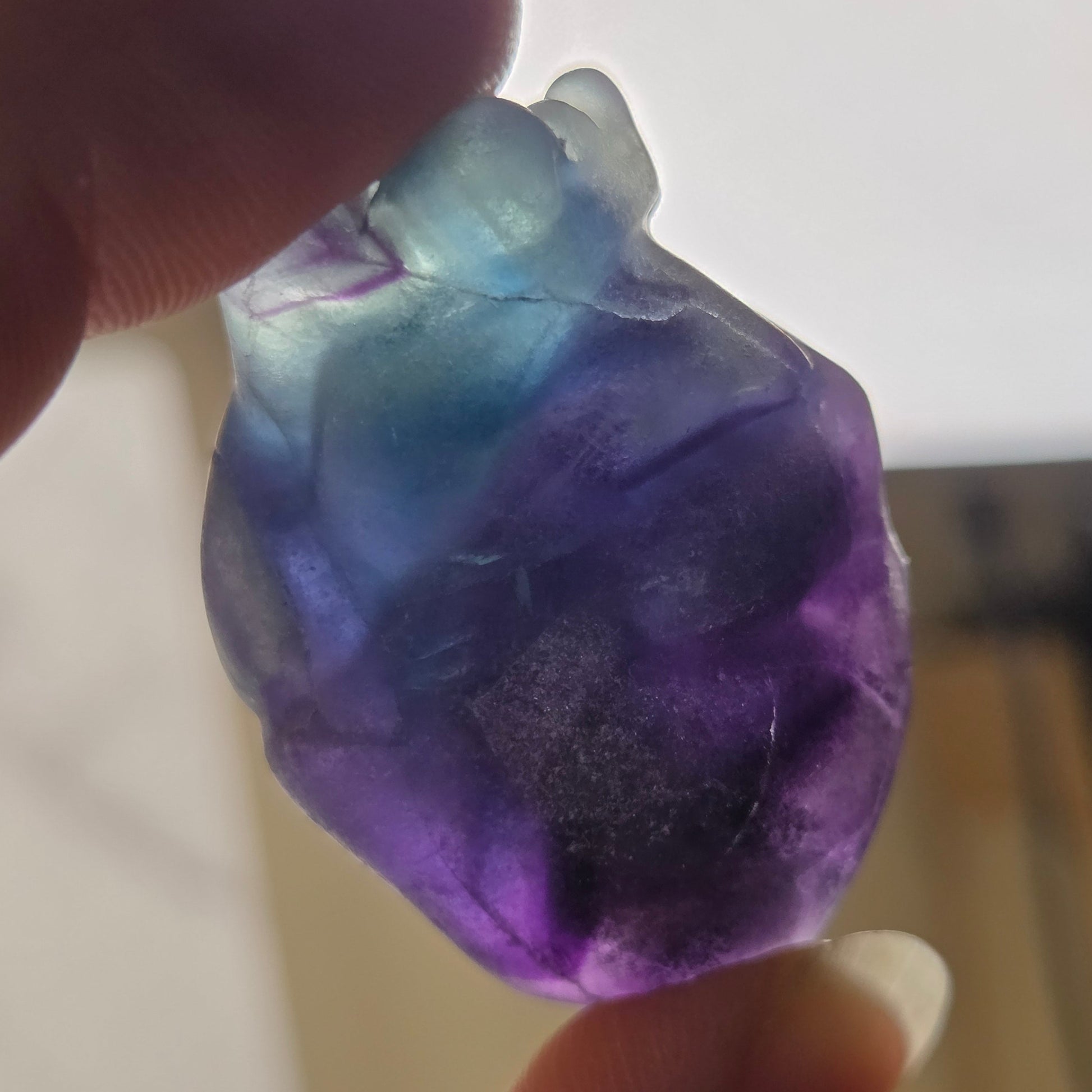 A hand holding a small, irregularly shaped translucent gemstone or crystal pendant with a color gradient from light blue at the top to deep purple at the bottom, photographed against a softly blurred indoor background.
