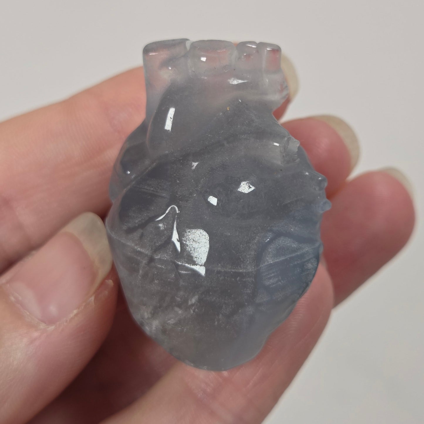 A translucent anatomical heart-shaped object, possibly a carved stone or resin piece, held in a person's hand against a plain light background.