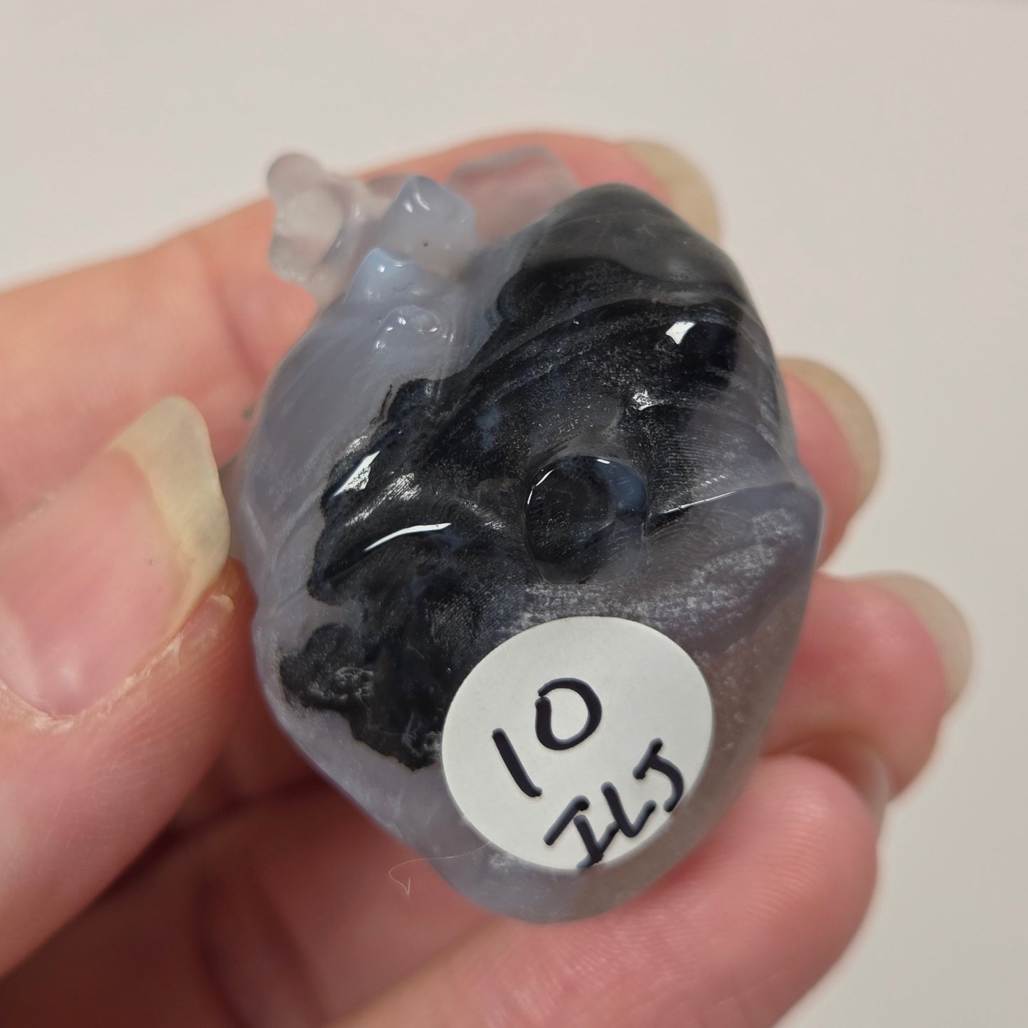 A hand holding a small, irregularly shaped translucent gray gemstone or crystal with a dark black inlay or embedded area. A white circular sticker with the handwritten numbers '10 215' is attached to the piece.