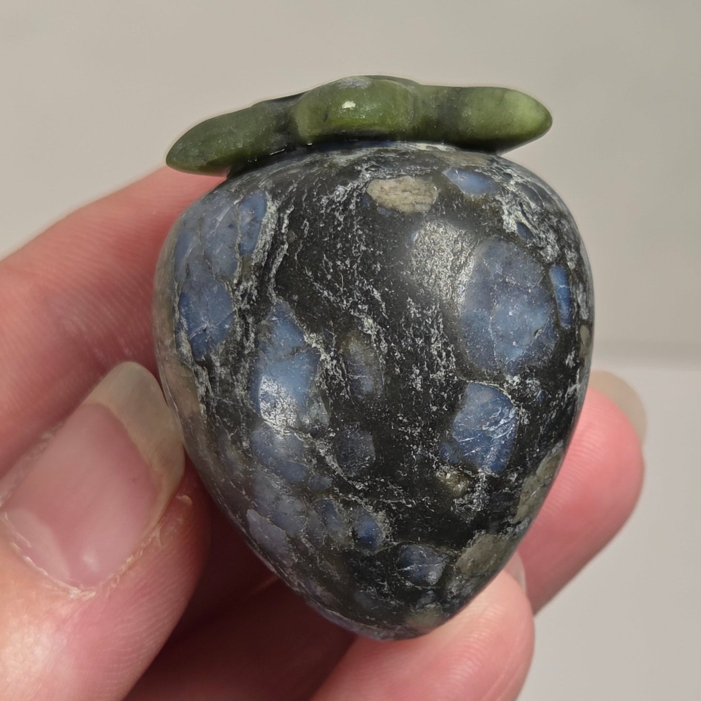 A small polished stone ornament shaped like a strawberry, held between fingers. The stone has a dark gray to black base with prominent light blue and white marbling, and a green stem at the top.