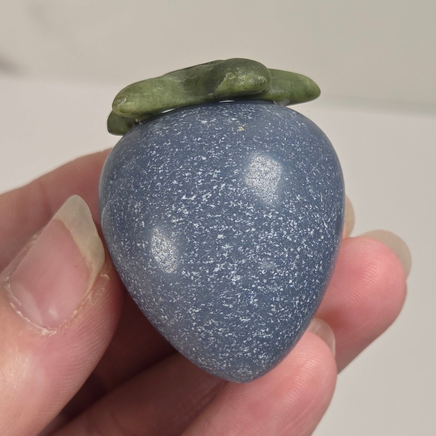 A hand holding a polished blue-gray stone fruit sculpture with a green leafy top, photographed against a light neutral background.