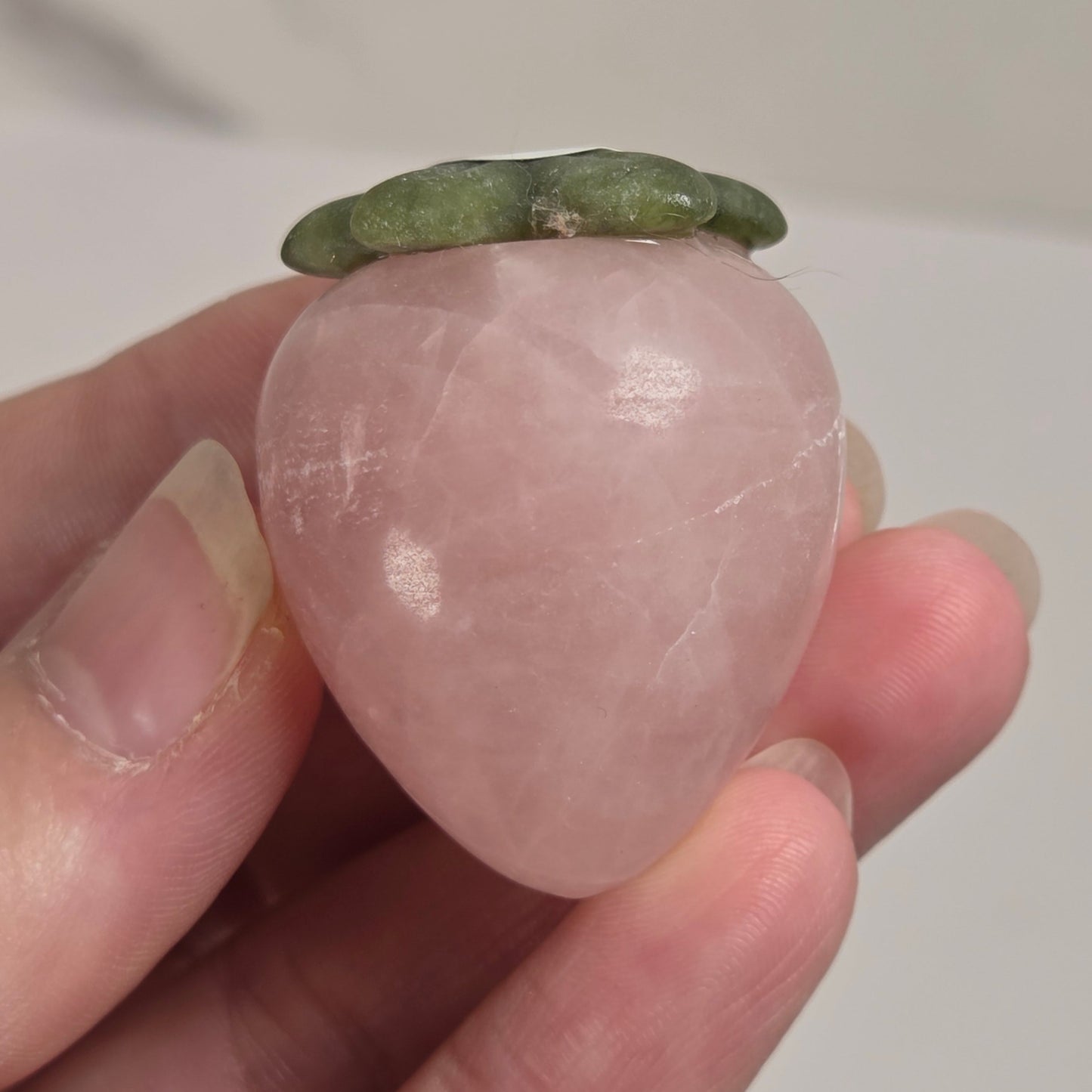 A hand holding a small peach-shaped rose quartz crystal with a green leafy top, shown against a light neutral background.