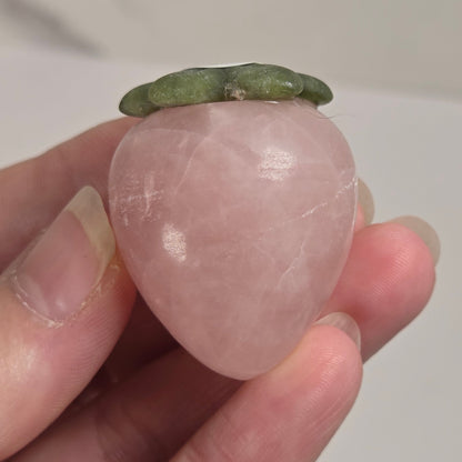 A hand holding a small peach-shaped rose quartz crystal with a green leafy top, shown against a light neutral background.