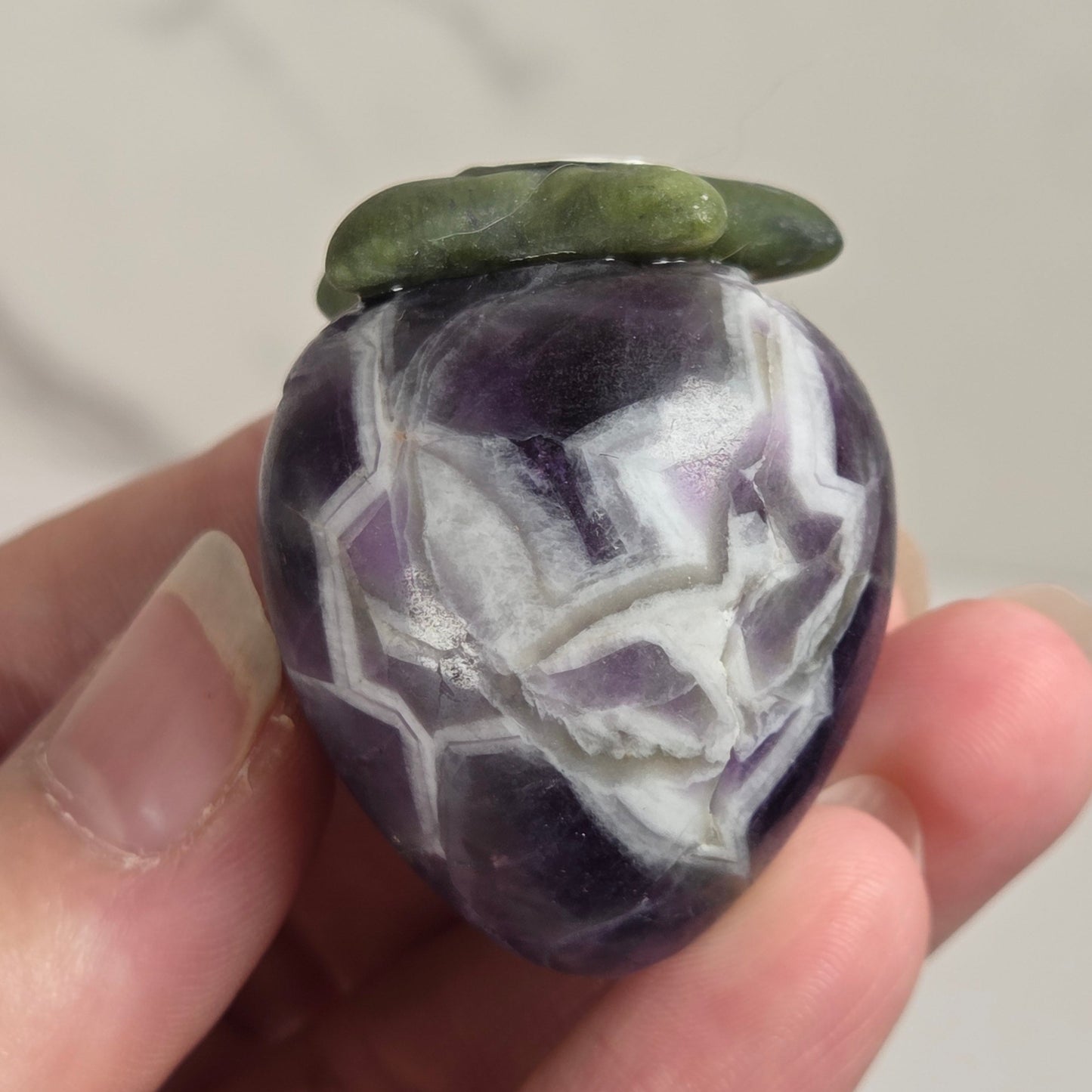 A hand holding a polished purple and white agate stone ornament with a green leaf-shaped top, set against a light marble background.