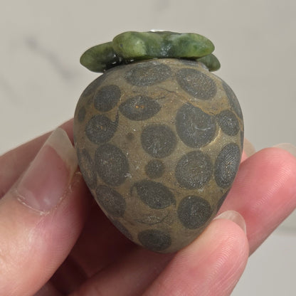 A hand holding a small decorative stone object shaped like a strawberry, with a mottled brown and gray body and green leaf-like top, against a light neutral background.