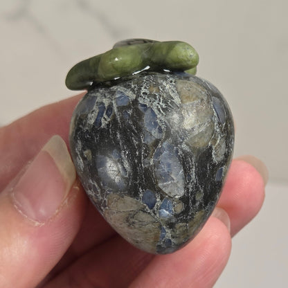A hand holding a polished labradorite stone ornament shaped like a pear, featuring natural gray and blue flecks with a green mossy top, set against a light marble background.
