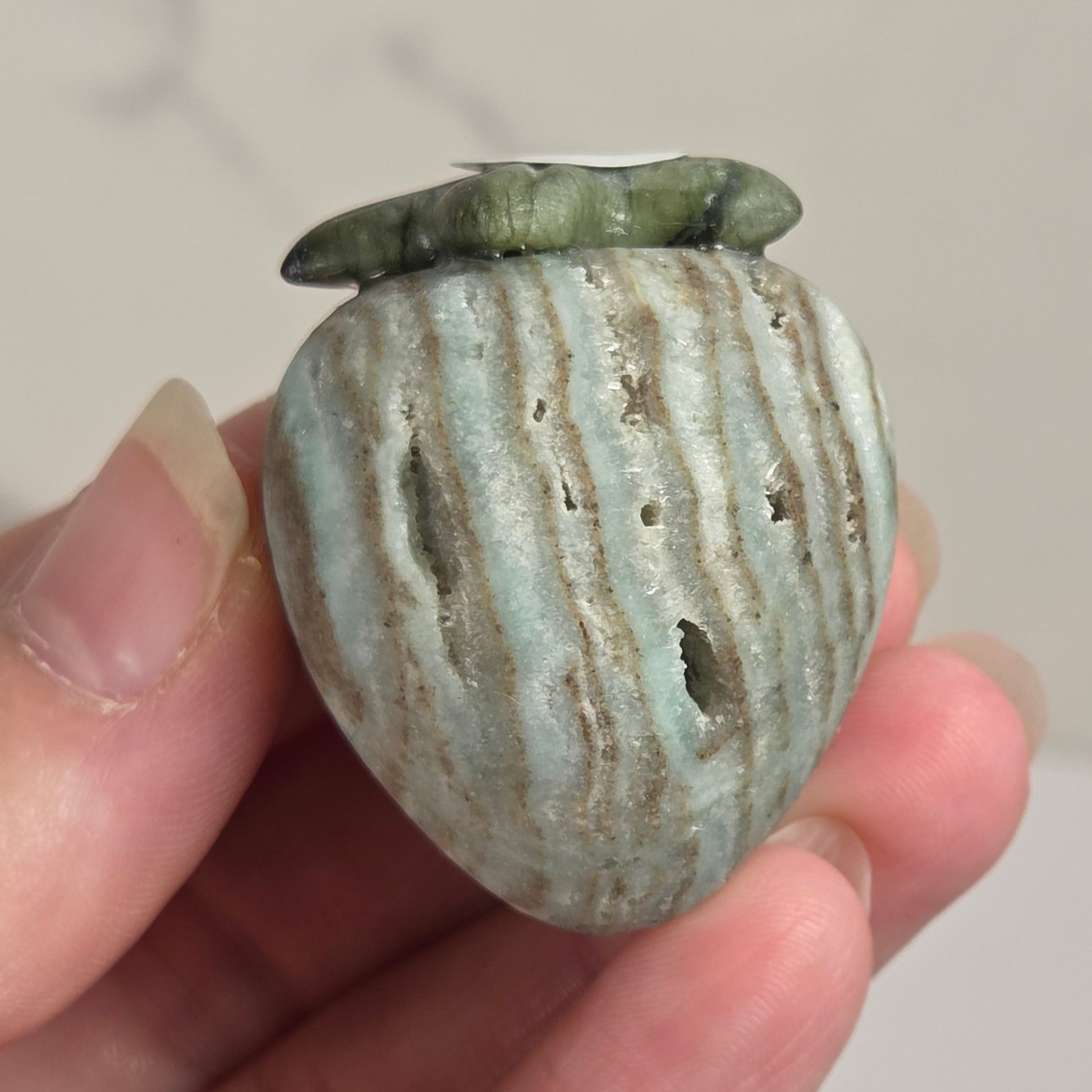A small decorative stone vessel with a rounded, ribbed body and a short flared top, held in a person's hand. The stone has a pale blue-green and beige striped pattern with darker brown veining.