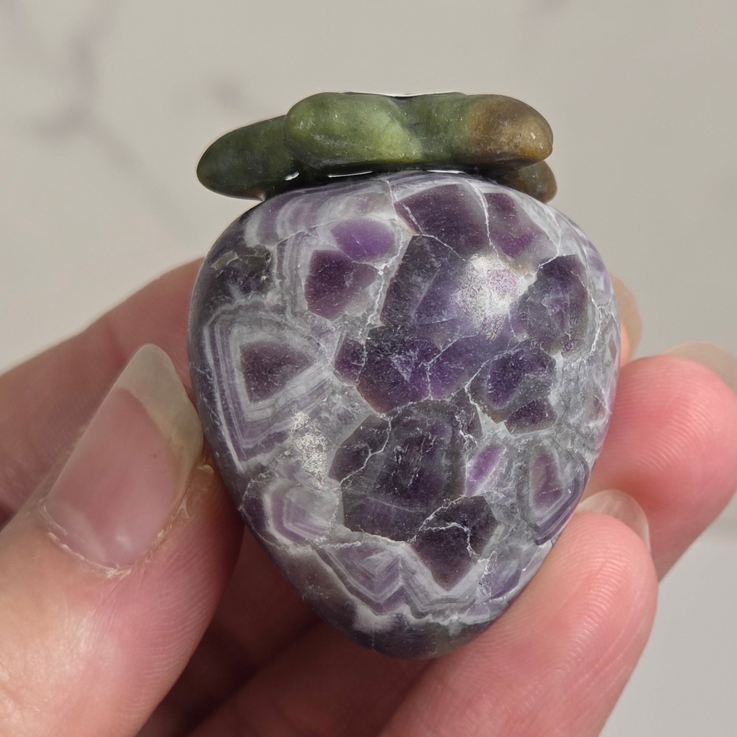 A hand holding a polished purple agate stone shaped like a strawberry, with green and brown moss accents on top, set against a light gray marble background.