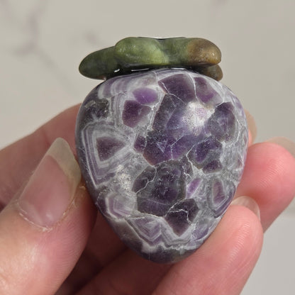 A hand holding a polished purple agate stone shaped like a strawberry, with green and brown moss accents on top, set against a light gray marble background.