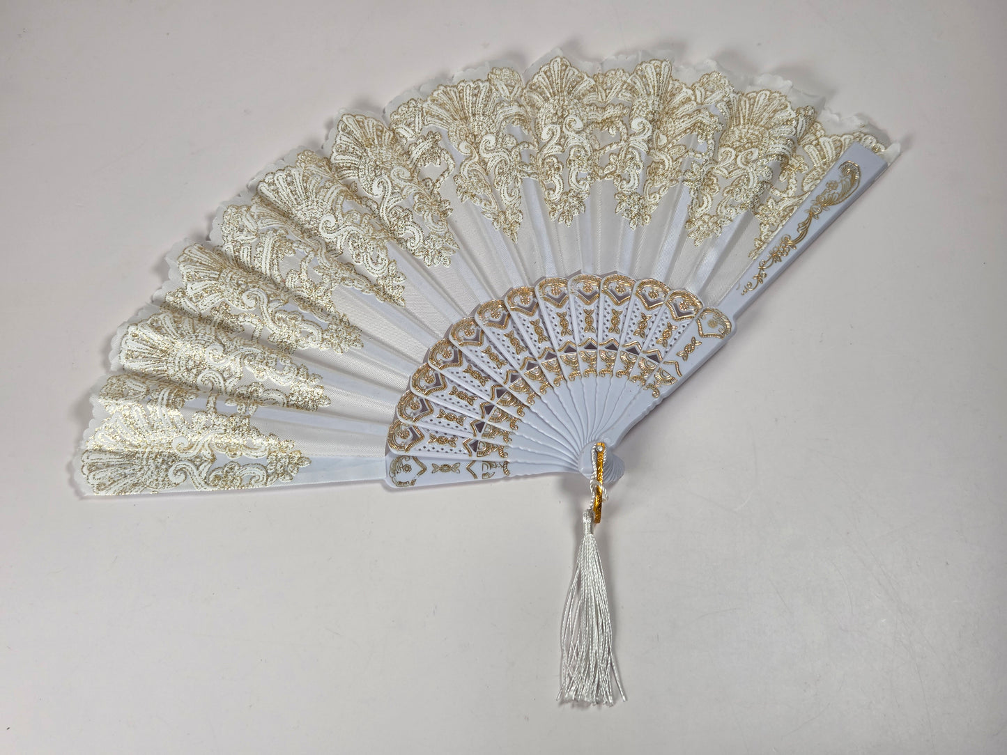 A white folding hand fan with intricate gold embroidered lace panels and a white tassel, displayed against a neutral light gray background.