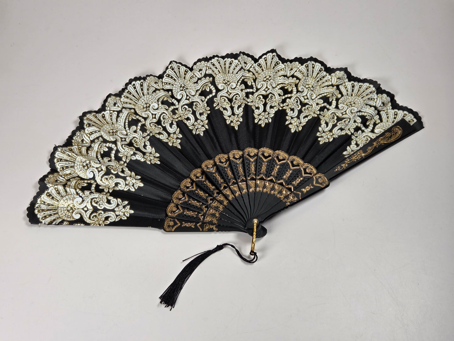 A black folding hand fan with ornate gold and white embroidered lace detailing on the upper section and a gold patterned lower section, featuring a black tassel.