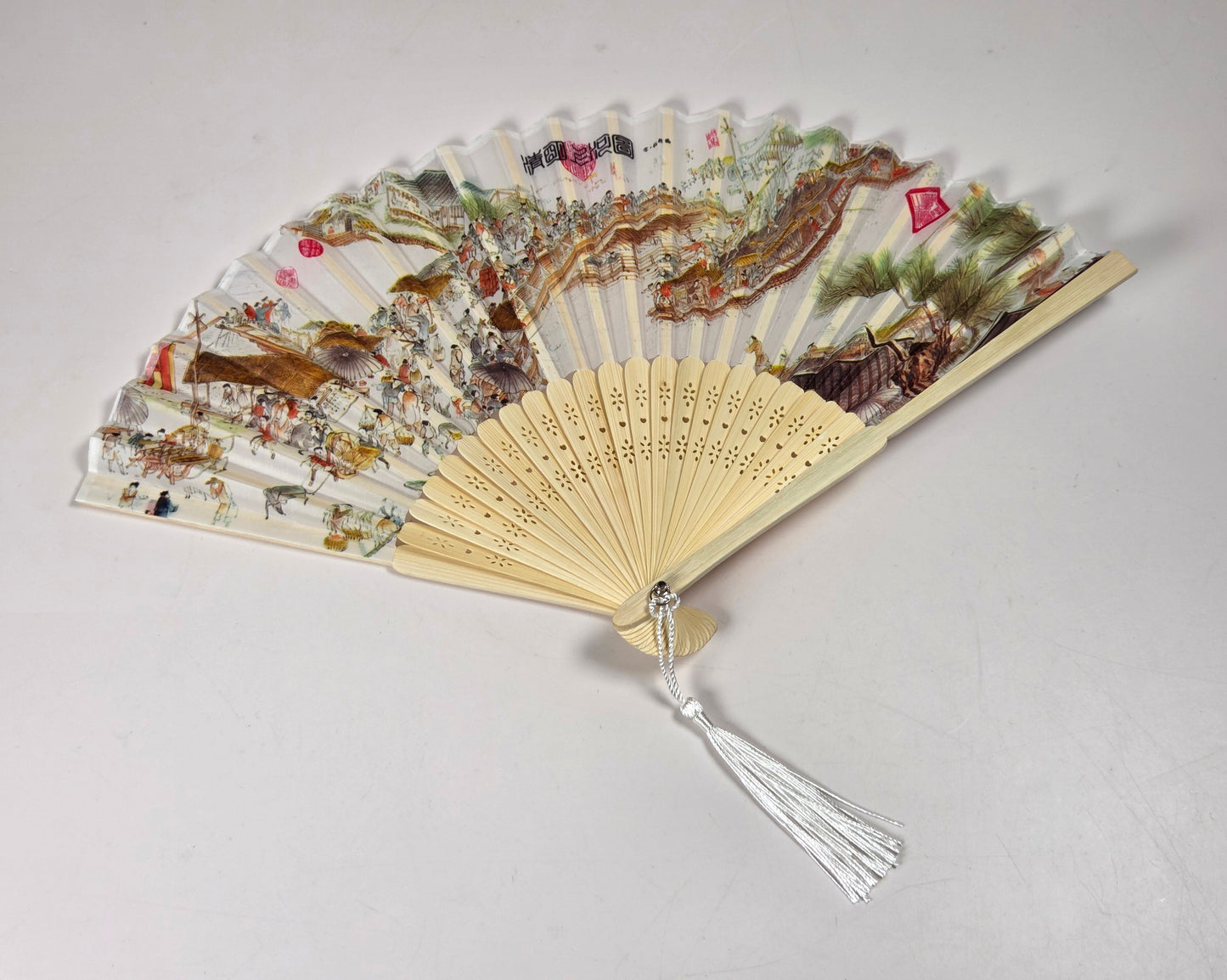 A traditional folding hand fan with a light beige bamboo frame, white tassel, and detailed multicolor printed paper panels depicting a scenic landscape with birds and foliage, shown on a plain light gray background.