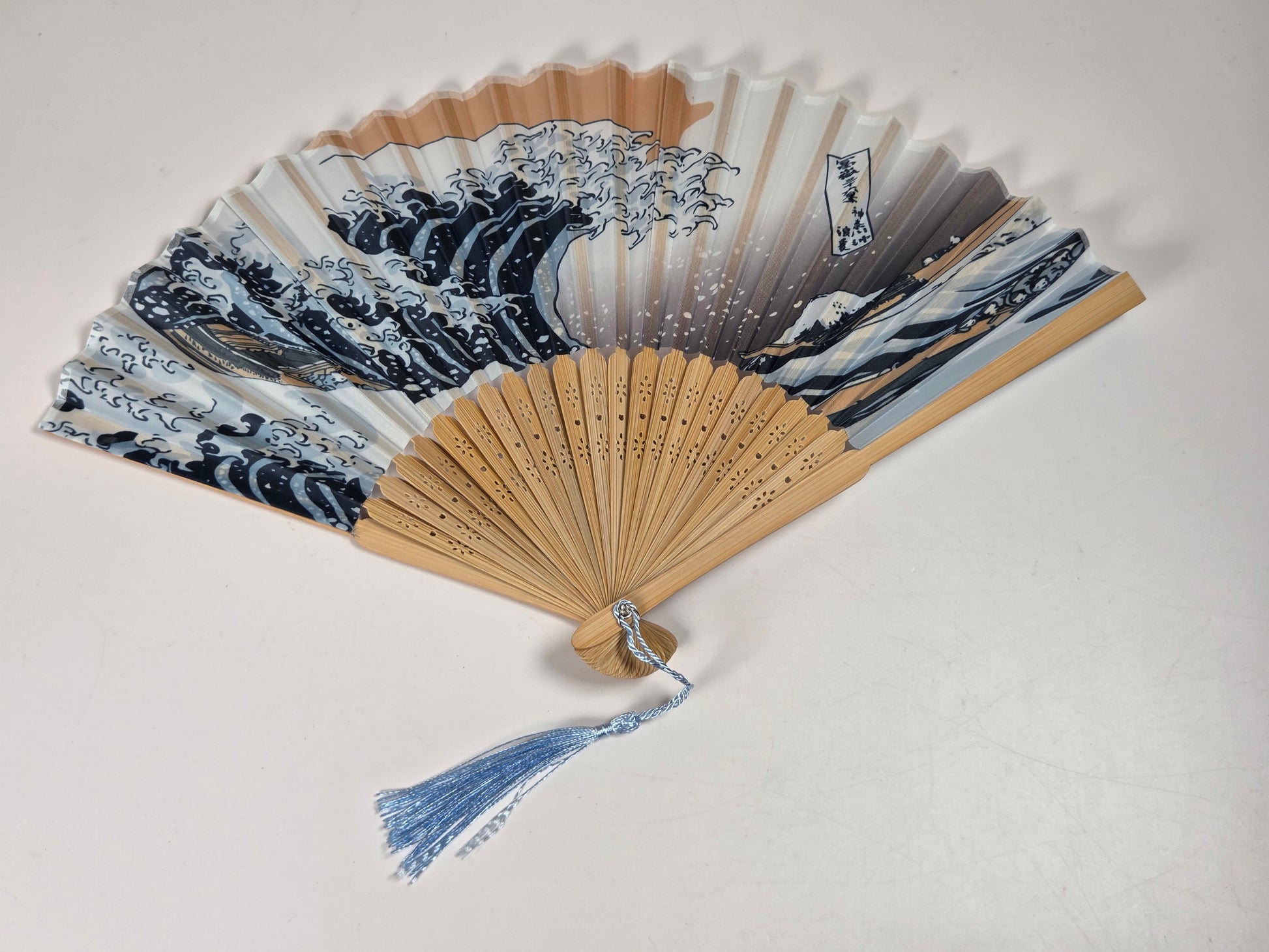 A folding hand fan with a light wood frame and a blue tassel, featuring a printed design of 'The Great Wave off Kanagawa' by Katsushika Hokusai. The wave pattern is in navy blue, white, and gray, while the wooden ribs are a natural light beige color.