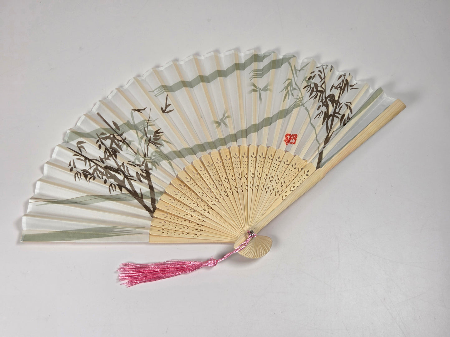 A traditional folding hand fan with a light bamboo frame, decorated with green and brown bamboo and leaf illustrations on the translucent white panels. It features a pink tassel at the top and a small red seal character on one panel, set against a plain light gray background.