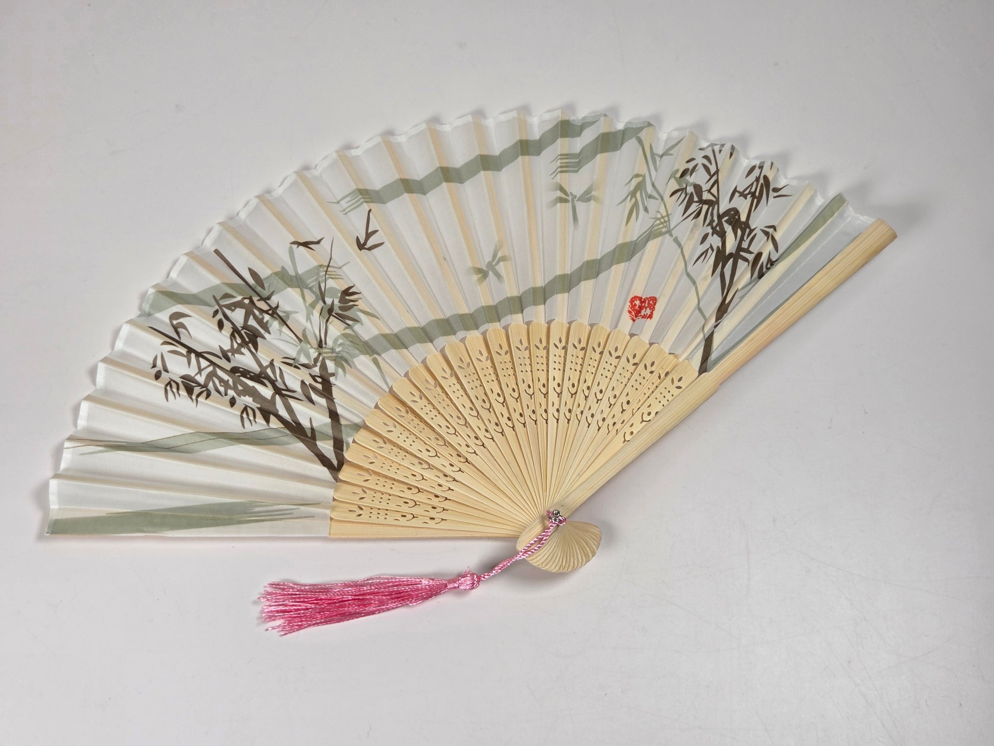 A traditional folding hand fan with a light bamboo frame, decorated with green and brown bamboo and leaf illustrations on the translucent white panels. It features a pink tassel at the top and a small red seal character on one panel, set against a plain light gray background.