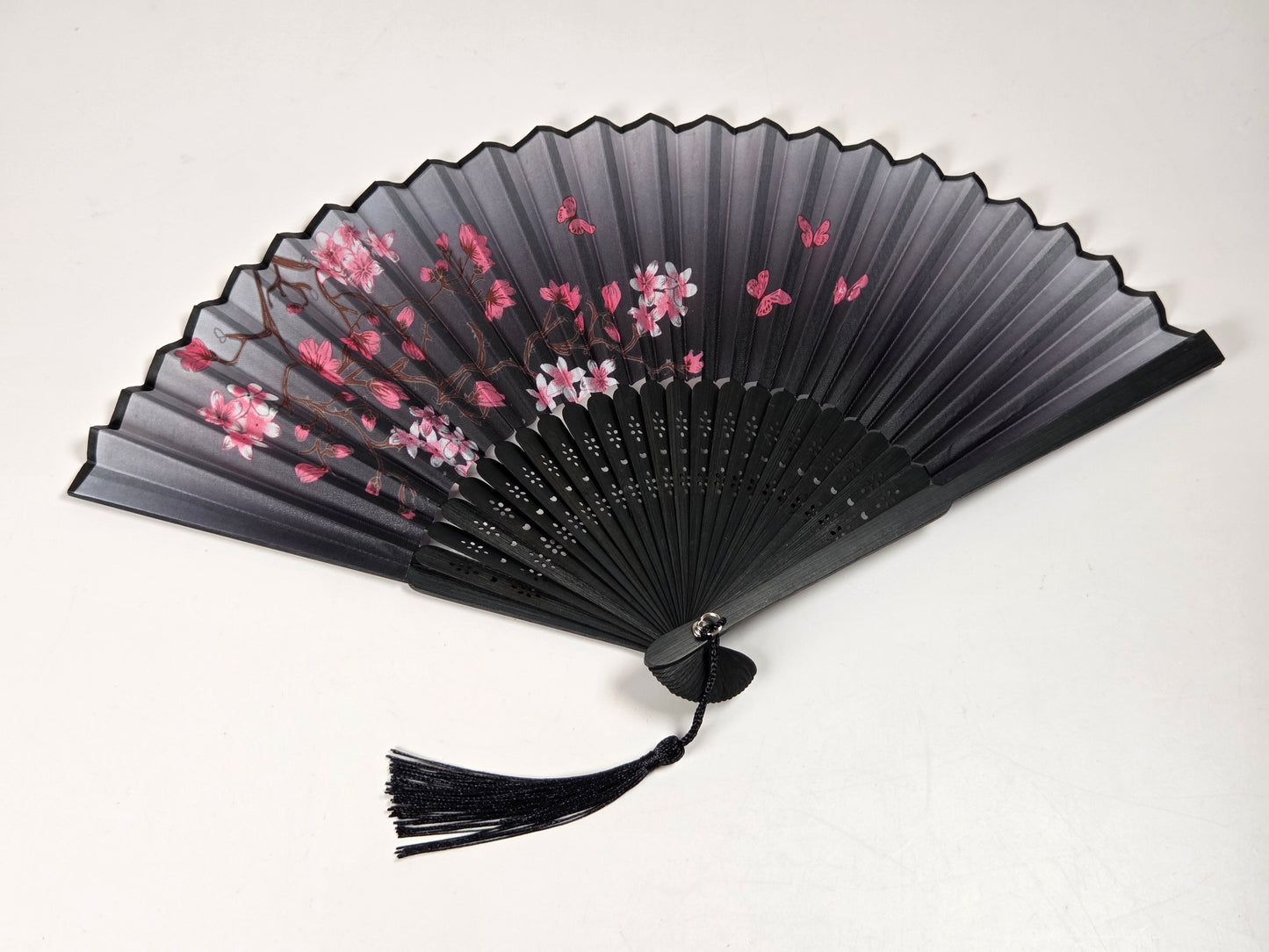 A black folding hand fan with a black tassel, decorated with pink and white cherry blossom flowers and brown branches, displayed against a plain off-white background.