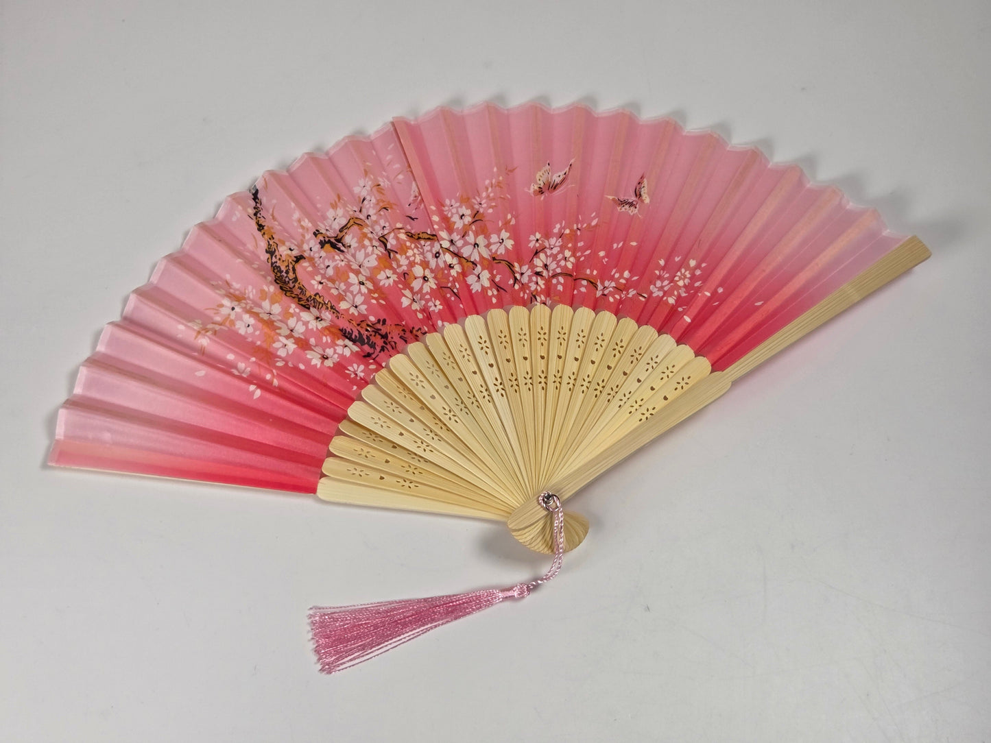 A traditional folding hand fan with a pink and peach gradient fabric, decorated with cherry blossom branches and fish motifs, featuring a pink tassel on a light background.