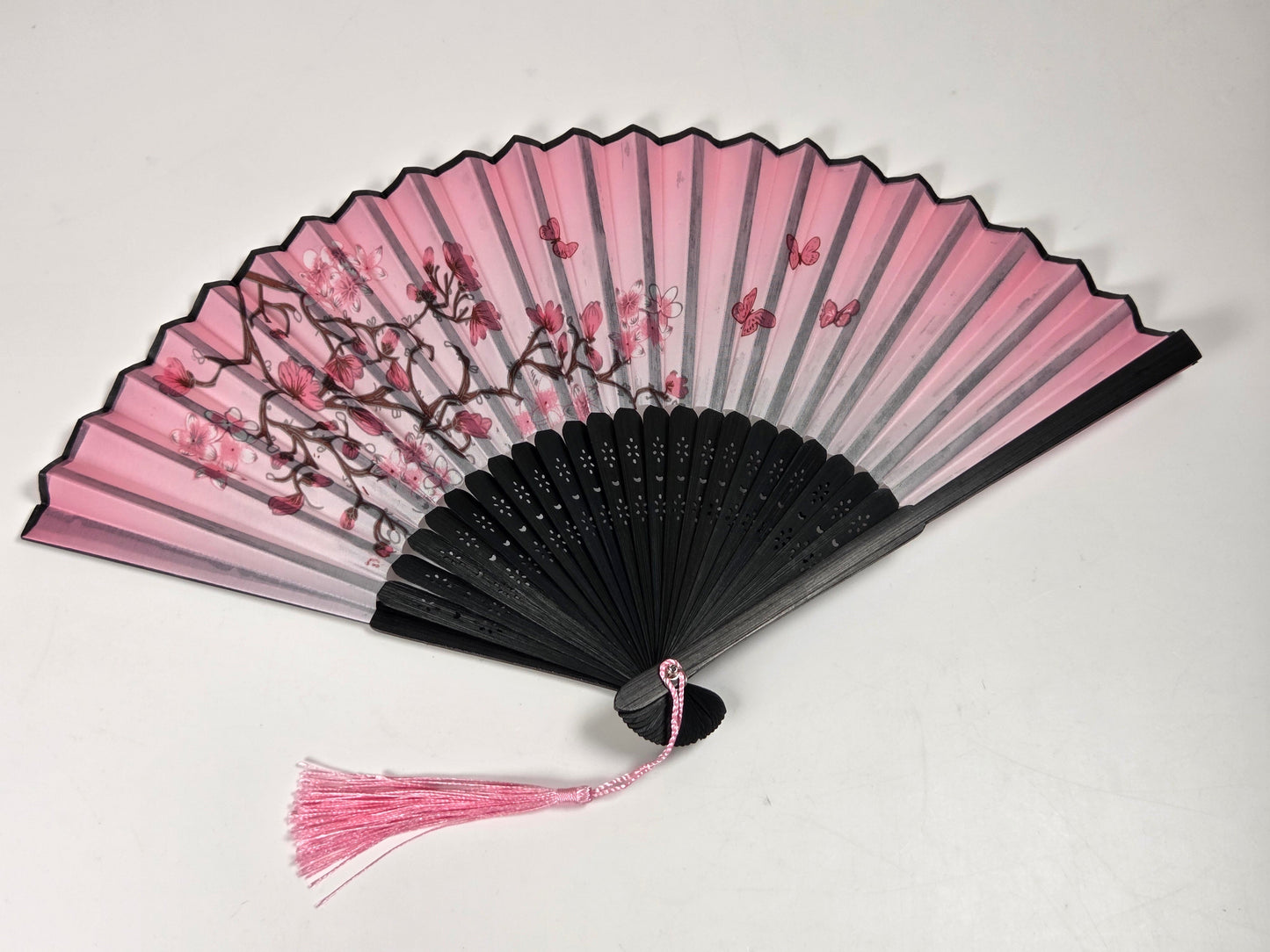 A traditional folding hand fan with a black and pink gradient design, decorated with cherry blossom branches and pink leaves, featuring a pink tassel on a light gray background.