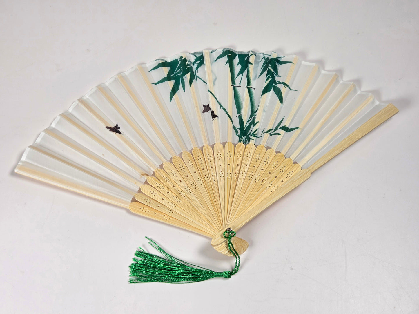 A traditional folding hand fan with a light beige bamboo frame, white paper panels decorated with green bamboo and small birds, and a green tassel at the top, shown on a plain off-white background.