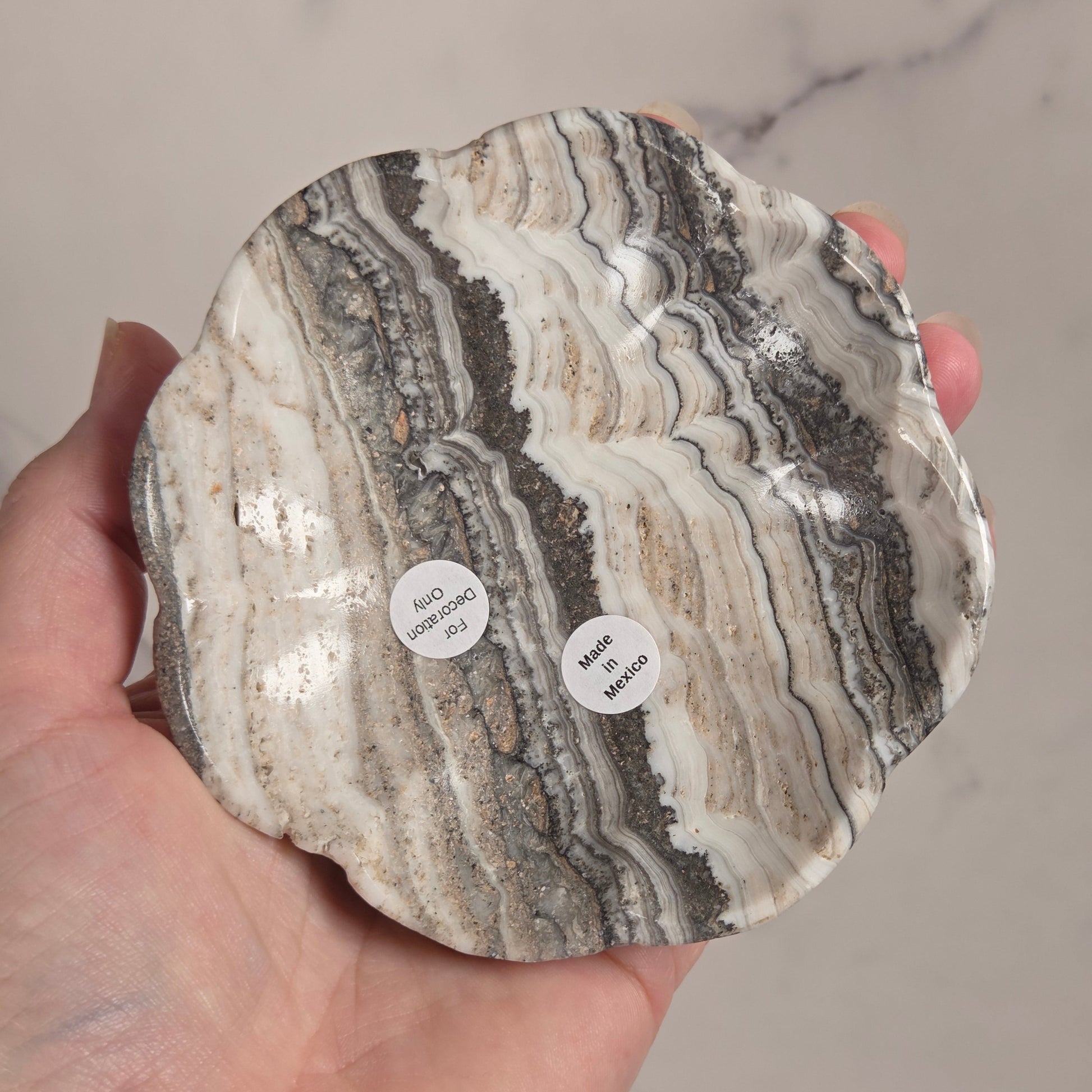 A hand holding a scalloped-edged stone dish made from layered agate or marble, showing wavy bands of cream, gray, brown, and black. Two small white circular labels are attached to the bottom of the dish.