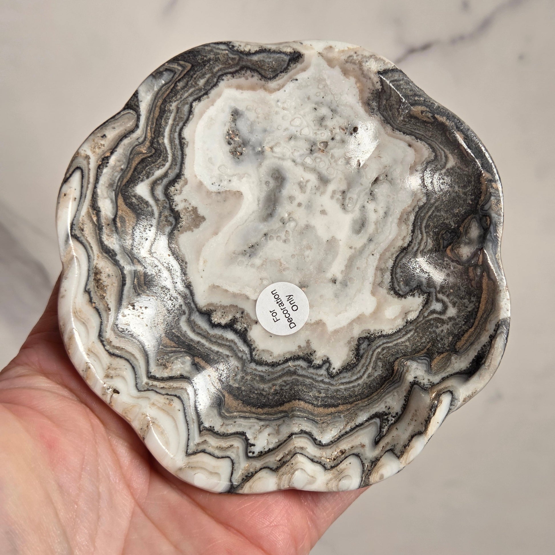 A hand holding a scalloped-edge ceramic plate or shallow dish made from polished agate or marble, showing wavy black, gray, and off-white natural stone patterns.