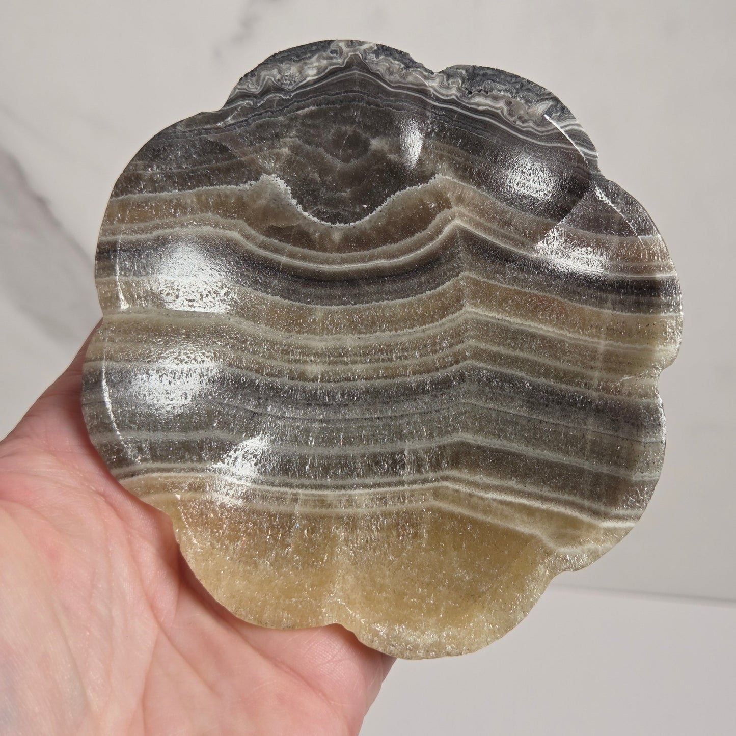 A hand holding a scalloped, flower-shaped stone tray made of layered agate or onyx with bands of gray, black, beige, and translucent golden-yellow.
