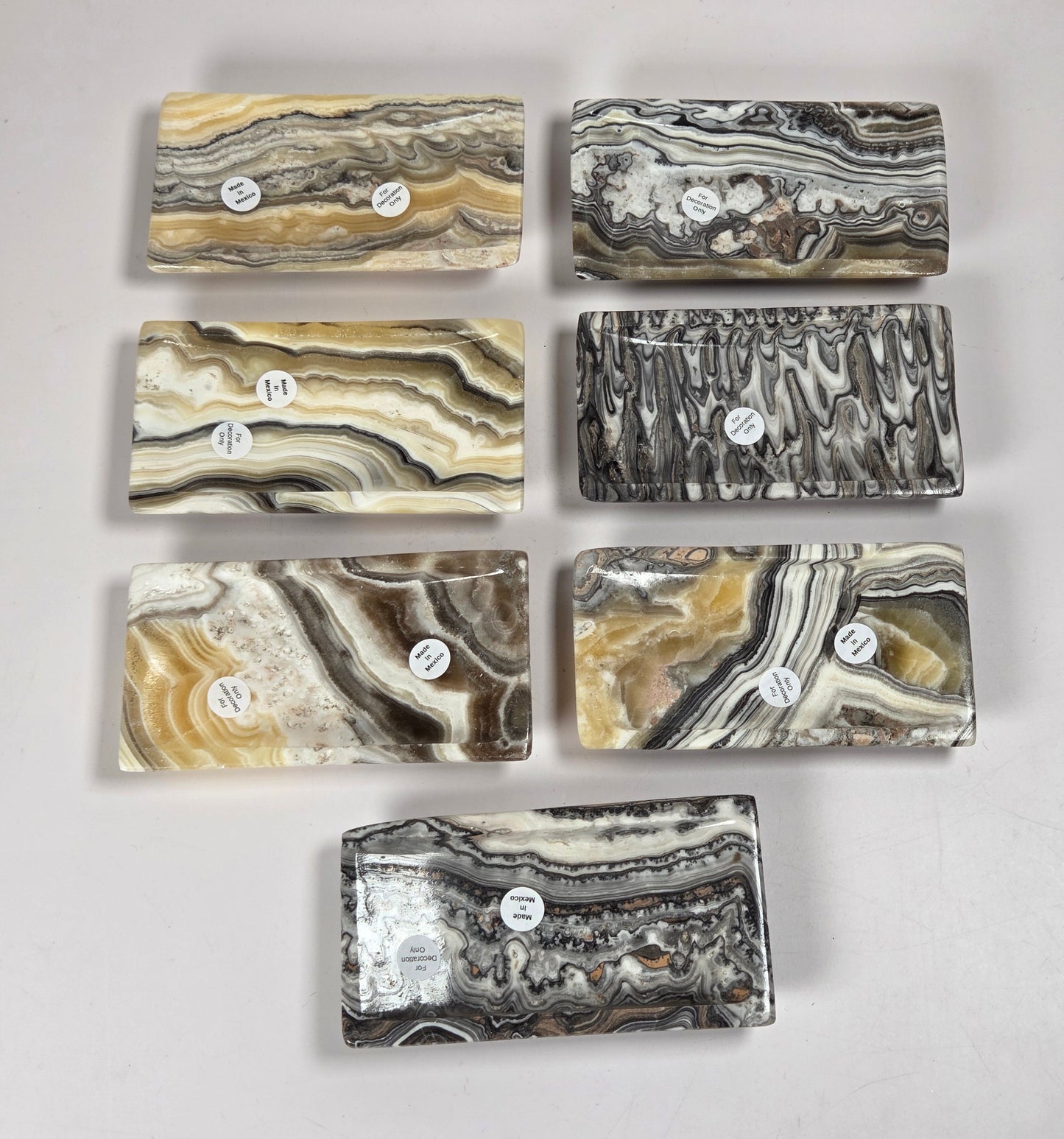 Six polished rectangular stone slabs with natural agate-like banding patterns in beige, gray, black, and brown, arranged on a light gray surface. Each slab has a small white circular label with handwritten numbers.