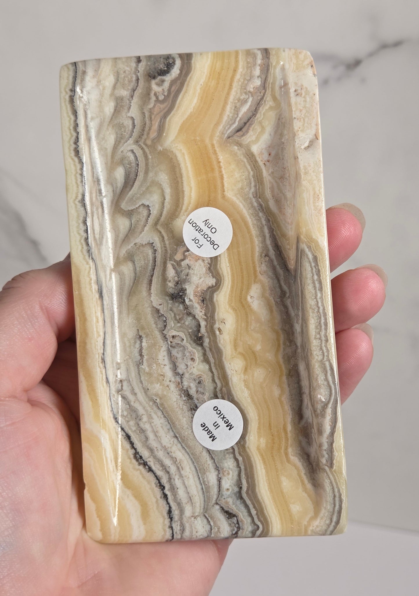 A hand holding a rectangular polished stone slab with wavy marbled bands of cream, tan, gray, and brown. Two small round stickers are attached to the front, and a light marble-patterned surface is visible in the background.
