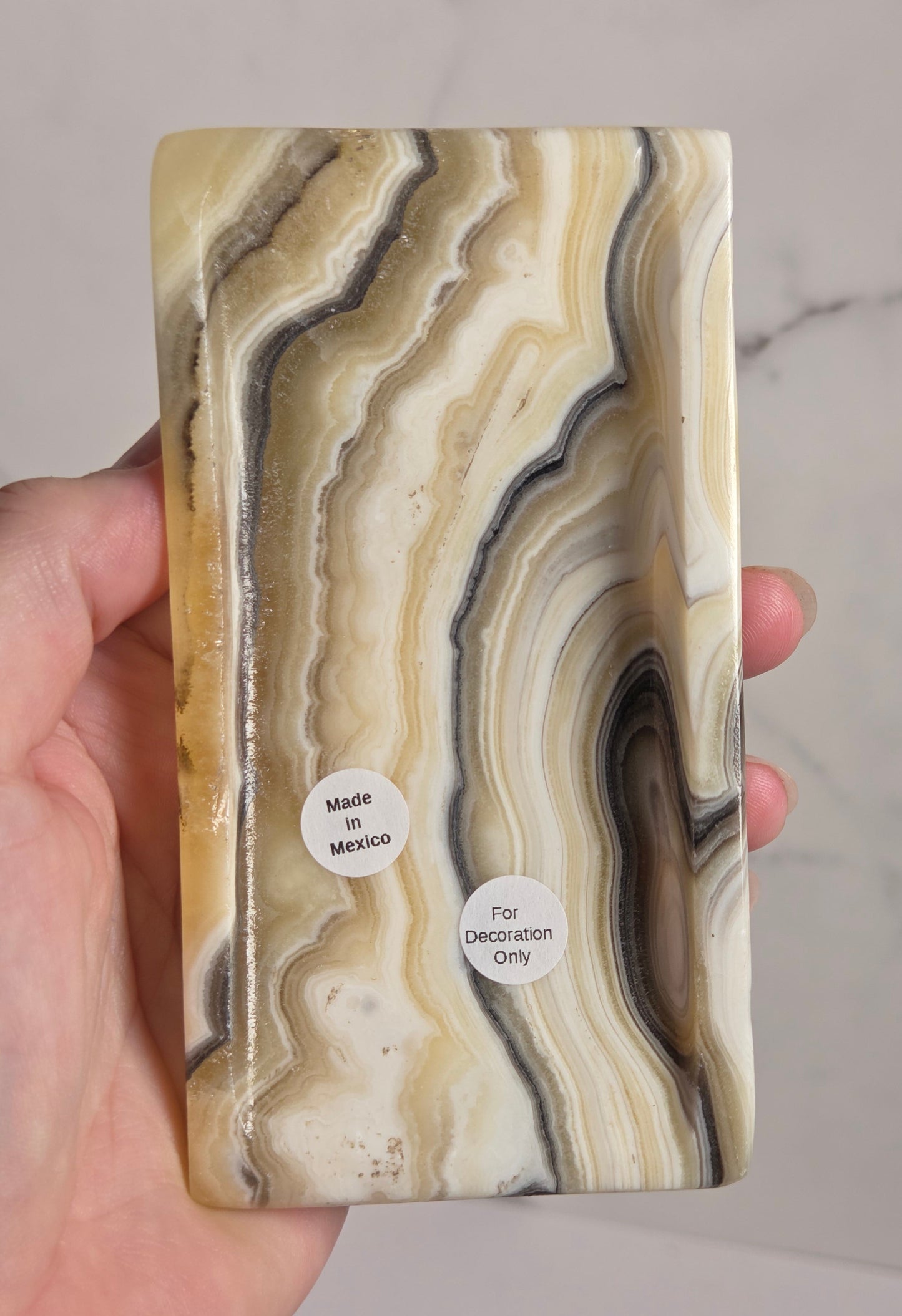A hand holding a rectangular resin plaque featuring a natural agate-like marbled pattern in cream, beige, brown, and gray tones, with two small white stickers reading 'Made in Mexico' and 'For Decoration Only'.