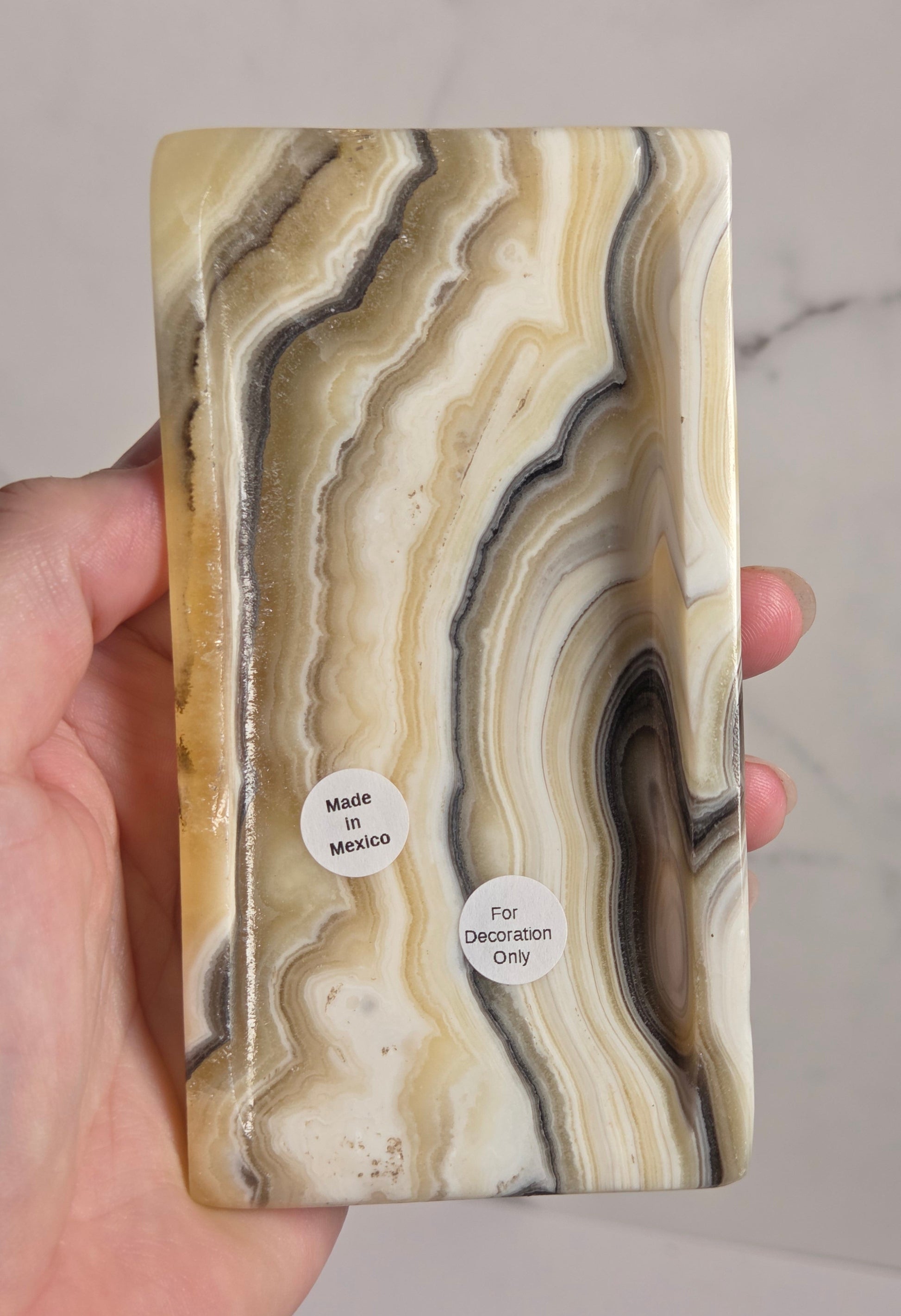 A hand holding a rectangular resin plaque featuring a natural agate-like marbled pattern in cream, beige, brown, and gray tones, with two small white stickers reading 'Made in Mexico' and 'For Decoration Only'.
