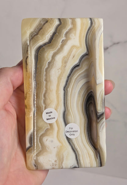 A hand holding a rectangular resin plaque featuring a natural agate-like marbled pattern in cream, beige, brown, and gray tones, with two small white stickers reading 'Made in Mexico' and 'For Decoration Only'.