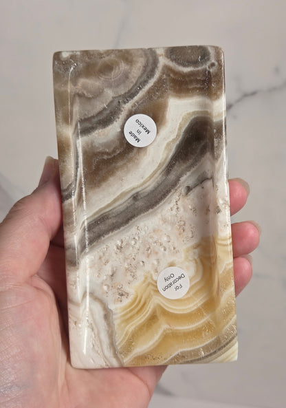 A hand holding a polished rectangular stone slab with swirling bands of cream, tan, brown, and gray, featuring small circular labels on the surface. The background is a light marble-patterned surface.