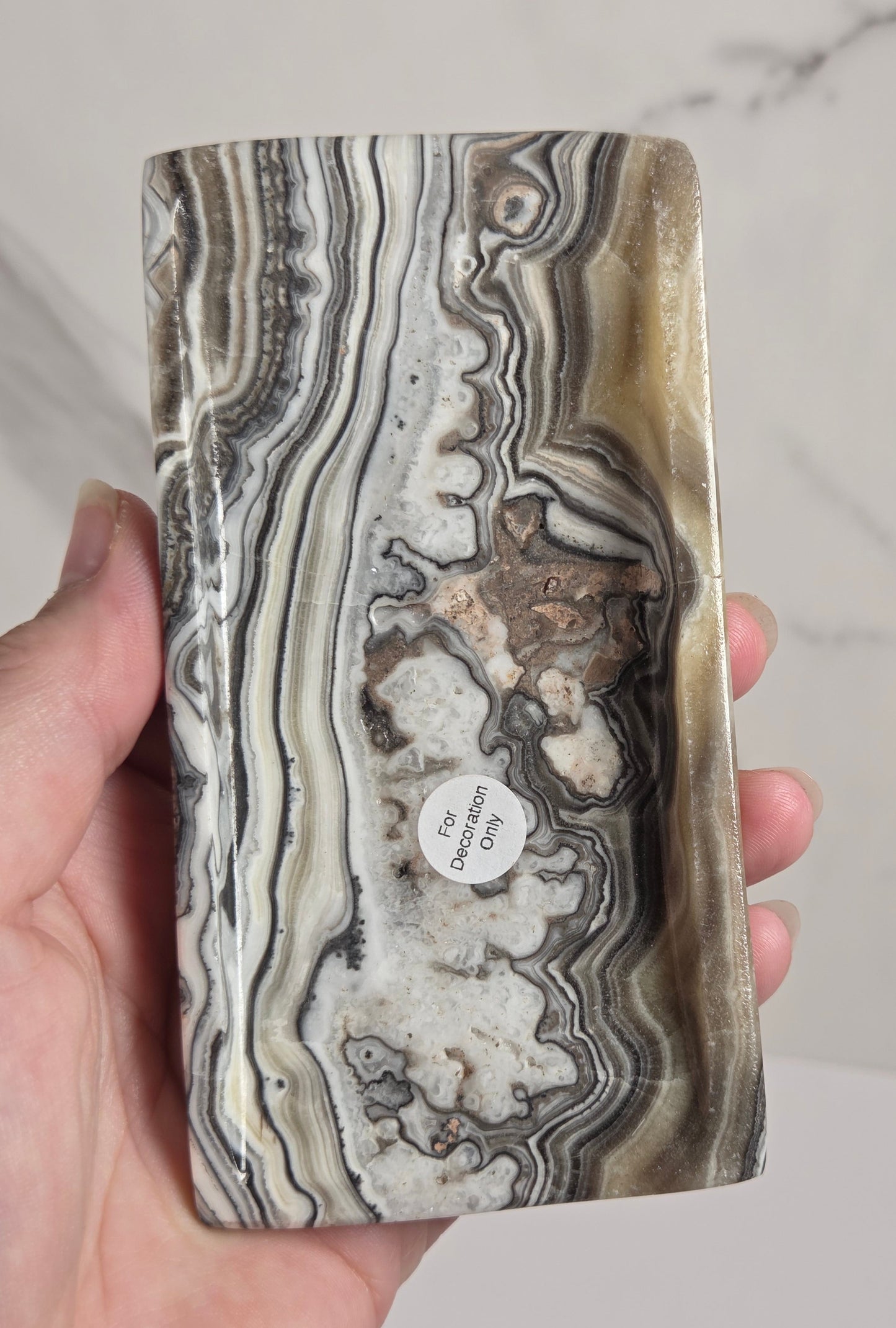 A hand holding a polished rectangular stone slab with swirling agate-like bands in gray, black, white, beige, and brown. A small round tag is attached near the lower center.