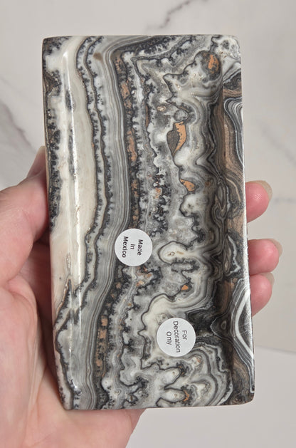 A hand holding a rectangular stone slab with swirling agate-like bands in gray, white, black, and brown, with two small white labels attached near the lower center.