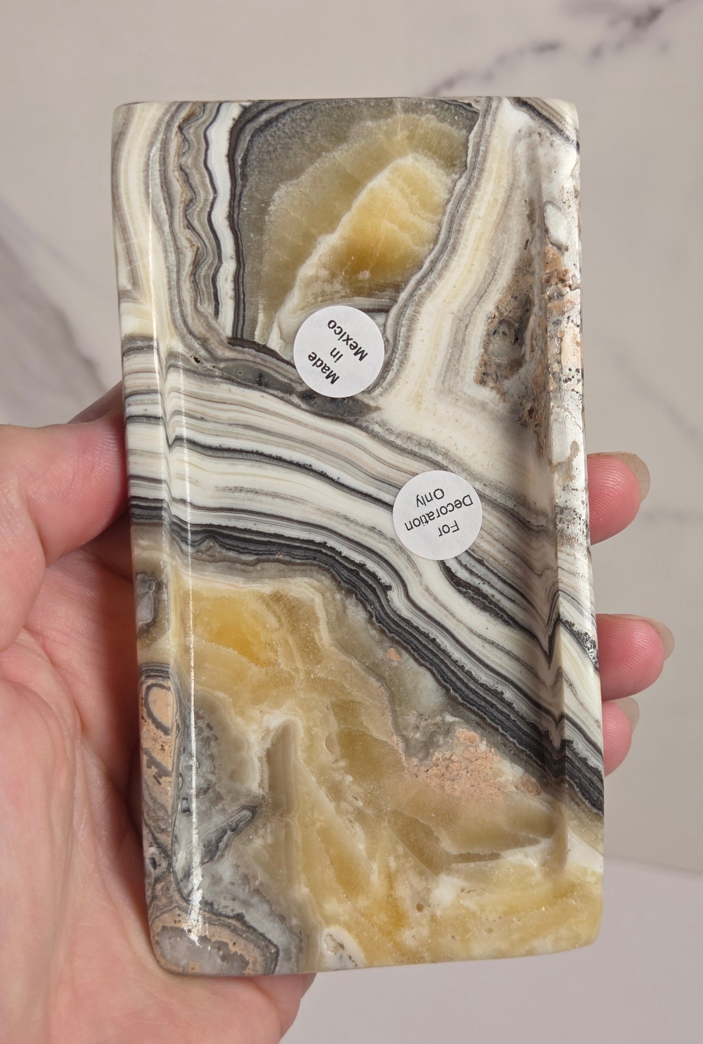A hand holding a rectangular polished stone tray with swirling bands of cream, beige, gray, black, and warm yellow tones. Two small white stickers are attached to the front, and a light marble-look surface is visible in the background.