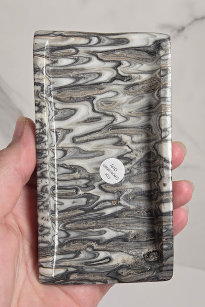 A hand holding a rectangular tray with a glossy black, gray, and white marbled pattern, set against a light marble background.