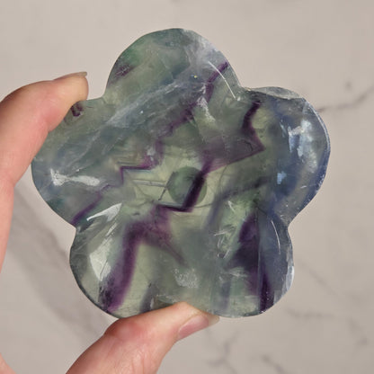 A hand holding a flower-shaped polished stone with marbled purple and green patterns, set against a light marble-patterned background.