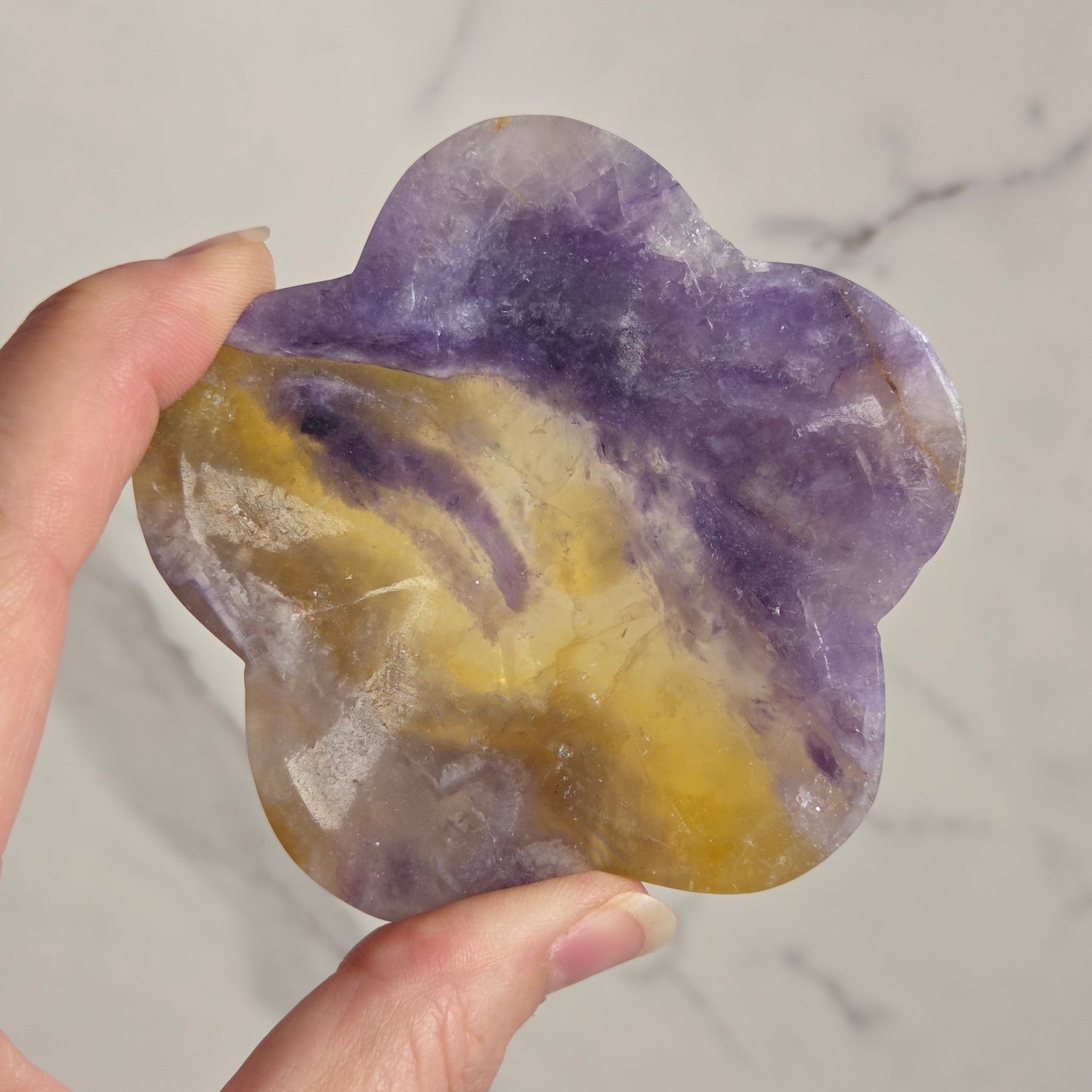 A hand holding a flower-shaped polished stone with marbled purple and yellow patterns, set against a light marble-patterned background.