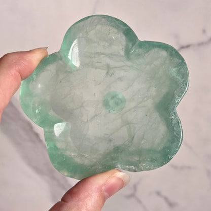A hand holding a flower-shaped polished stone made of green fluorite, set against a light marble-patterned background.