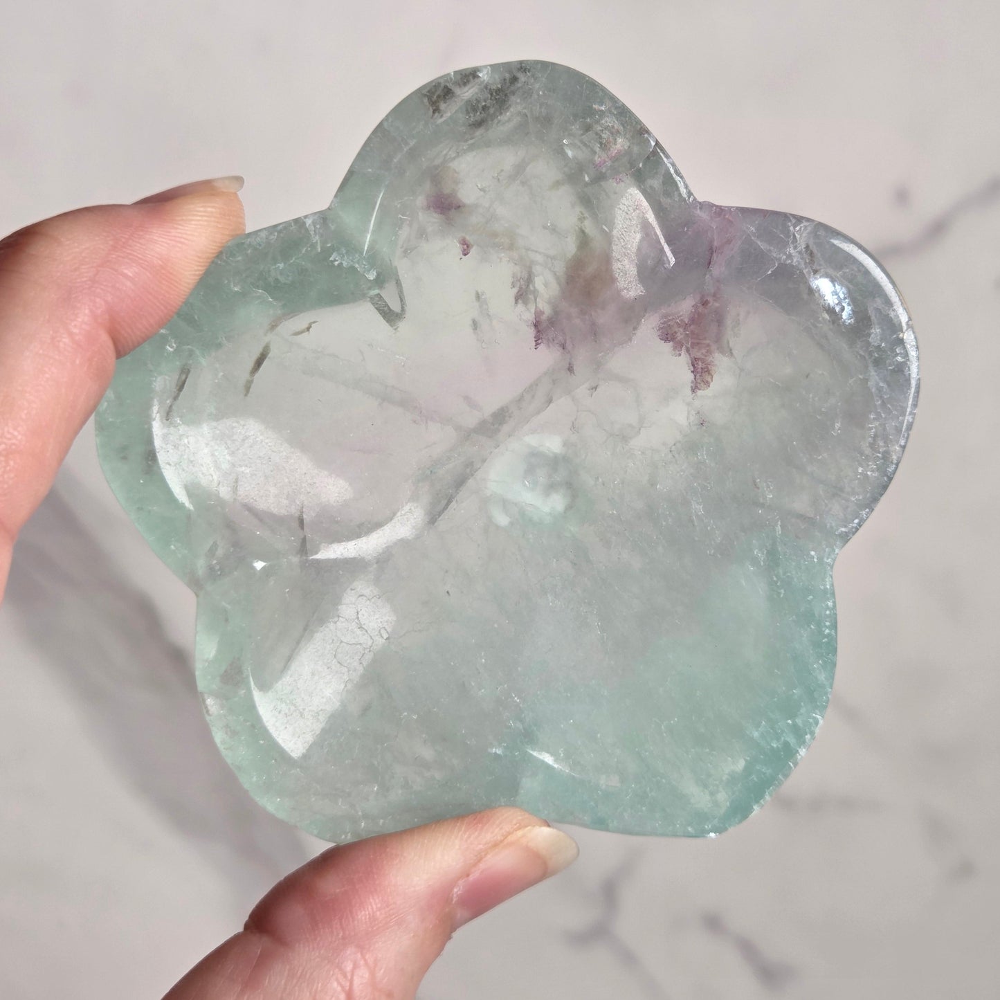 A hand holding a flower-shaped polished stone made of green fluorite, set against a light marble-patterned background.