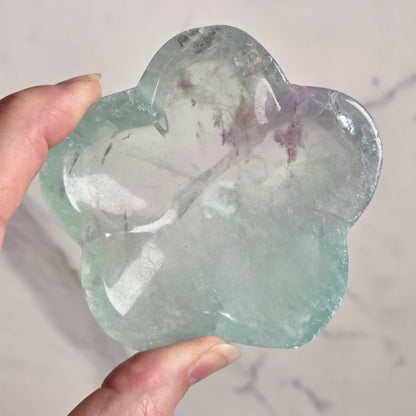 A hand holding a flower-shaped polished stone made of green fluorite, set against a light marble-patterned background.