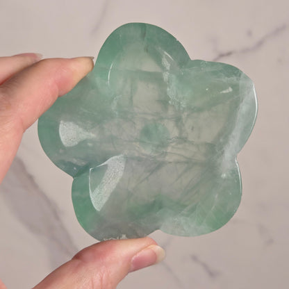 A hand holding a translucent light green carved stone piece with a rounded, organic shape against a light marble-patterned background.