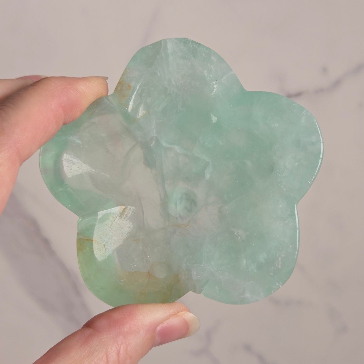 A hand holding a carved light green stone ornament shaped like a flower, shown against a pale marble-patterned background.