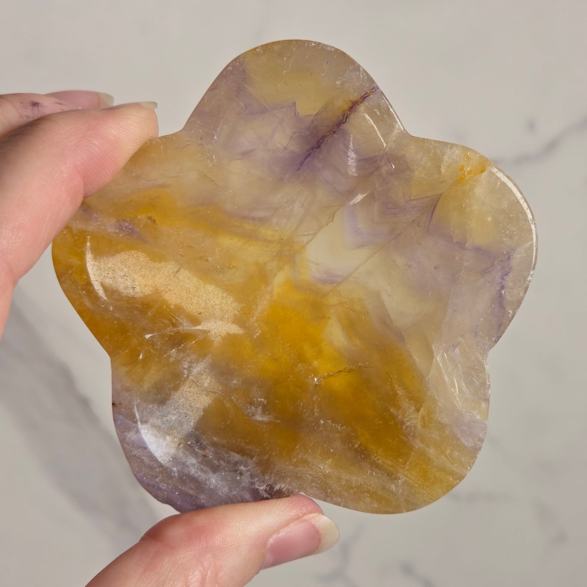 A hand holding a polished, flower-shaped crystal stone with marbled yellow, orange, and purple hues against a light gray marble background.