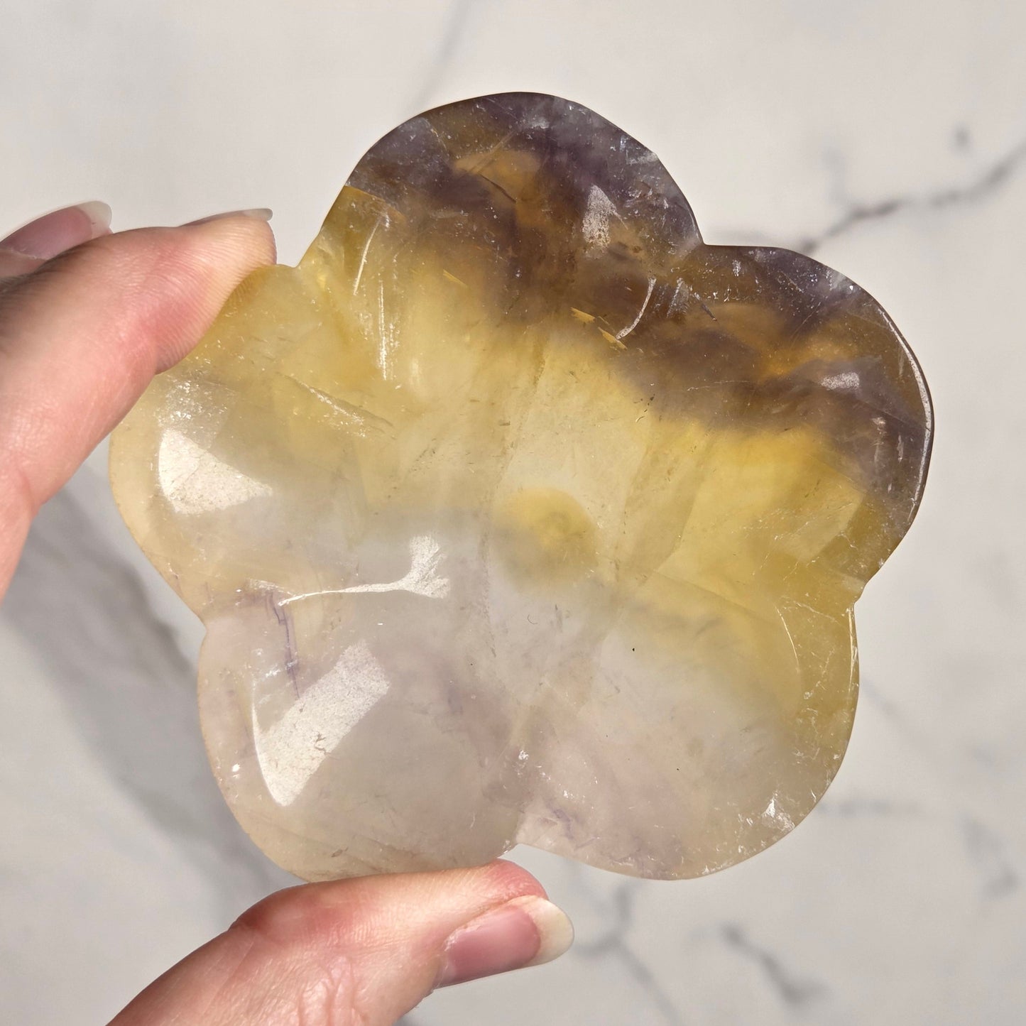 A hand holding a polished, flower-shaped agate crystal with natural banding of yellow, brown, and purple hues, set against a light gray marble background.