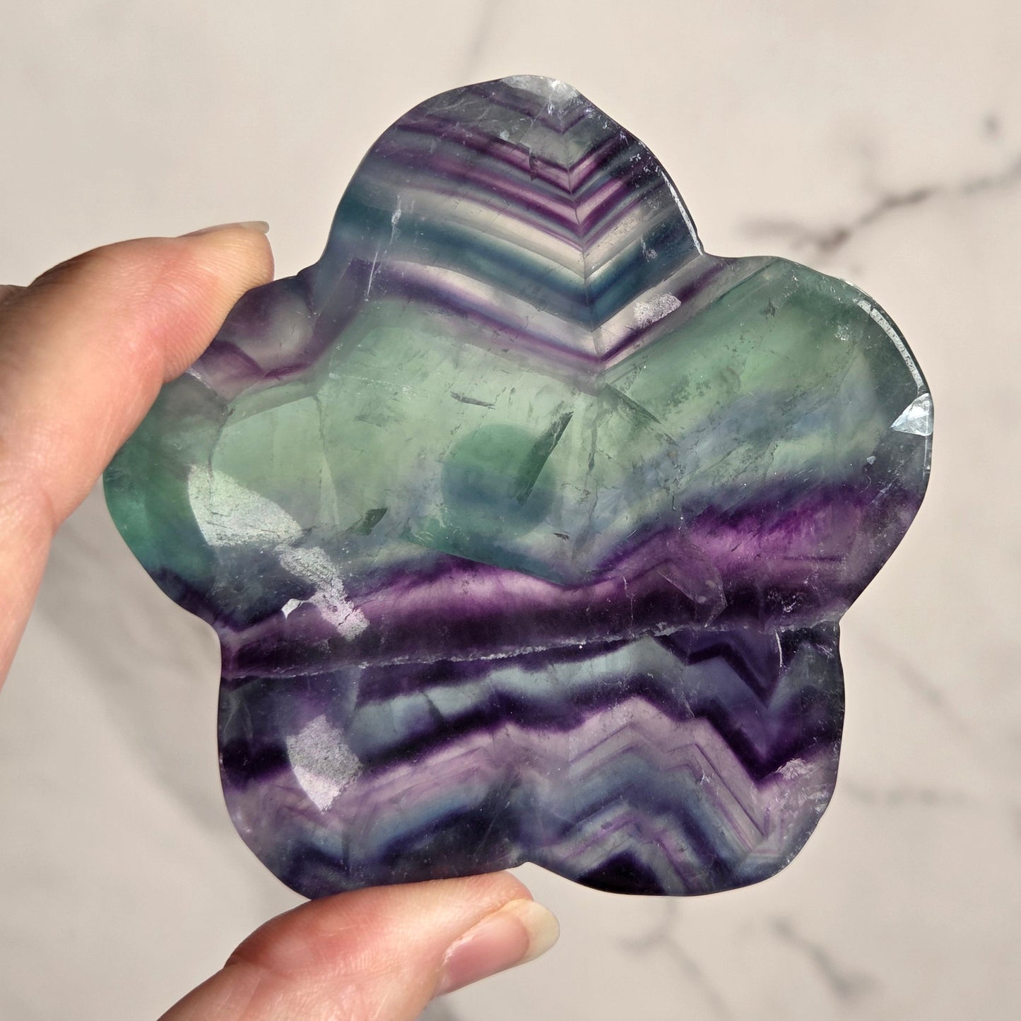 A hand holding a flower-shaped polished stone with layered bands of green, purple, and dark gray, set against a light marble background.