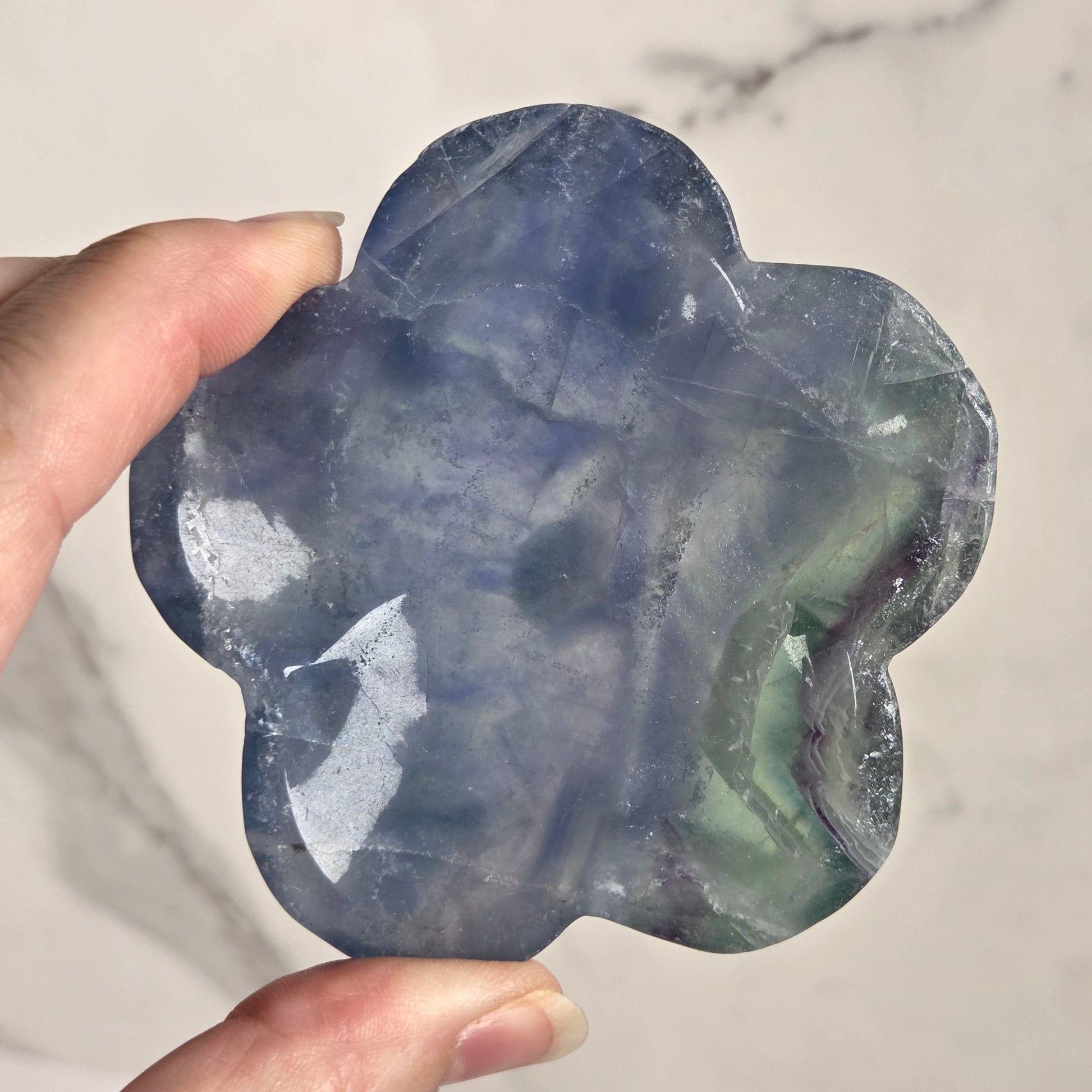 A hand holding a polished, flower-shaped blue stone with green and purple inclusions, set against a light marble background.