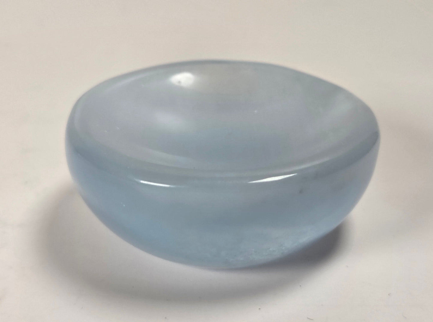 A small, smooth, dome-shaped translucent light blue glass object, possibly a dish, candle holder, or decorative piece, shown on a plain off-white surface.