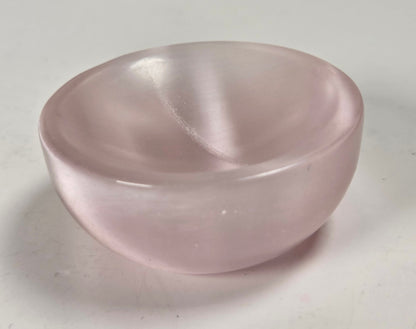 A small, smooth, semi-translucent pink stone bowl, likely rose quartz, photographed on a plain light gray surface.