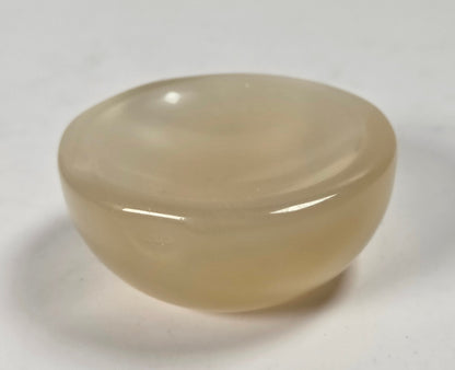 A small, smooth, rounded translucent stone object with a warm beige-tan hue, photographed on a plain light background.